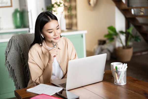 young asian woman studying student listening webinar from home wearing earphones looking lapt